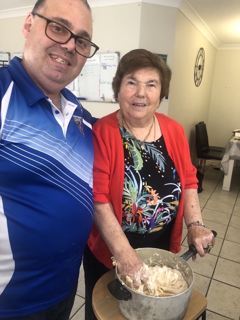A man and an elderly woman standing in a kitchen, with the woman mixing dough in a large pot.